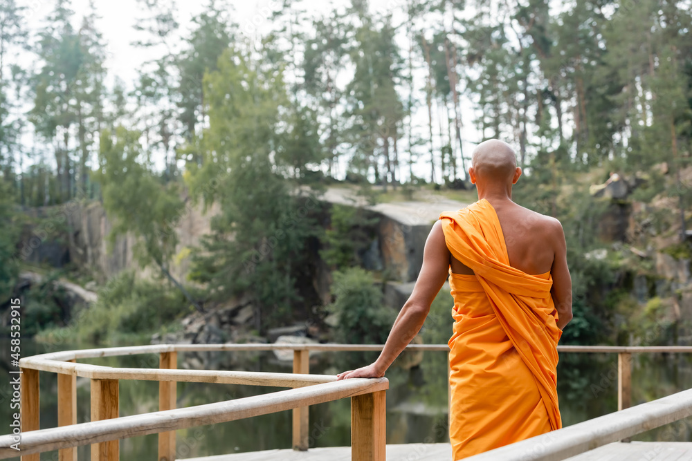 back view of buddhist meditating near wooden fence over lake in forest