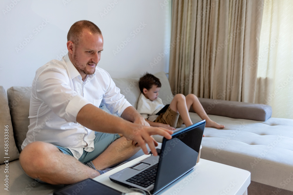 Attractive mature dad with adjust to the new situation due to Coronavirus issue, started working from home and taking care of his son. Young businessman working on laptop from home.