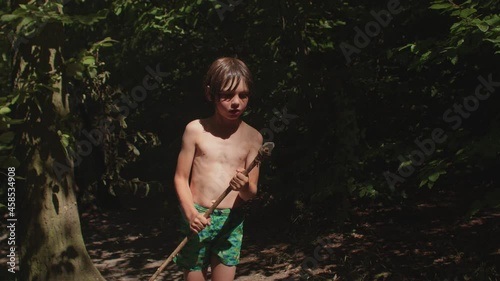 Portrait of a boy with a handmade spear in the forest. Long haired preteen playing survival game outdoor. The stone tip is tied with a rope. The child is wearing green swim shorts.