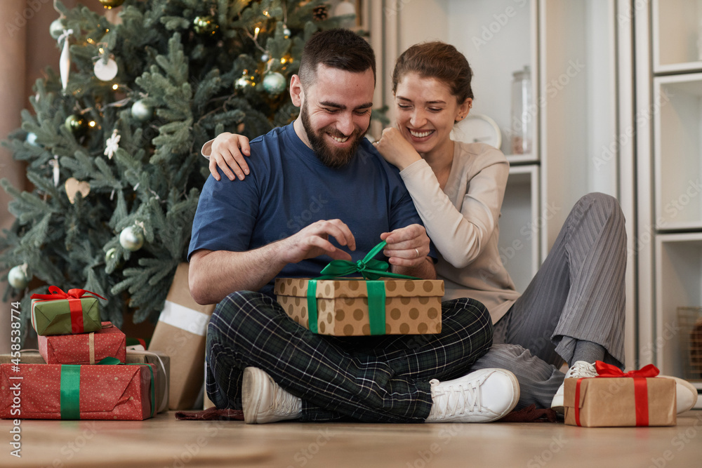 Smiling young bearded man embraced by girlfriend sitting with crossed legs against Christmas tree and unwrapping present