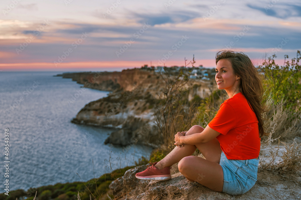 A young woman tourist stands on the shore high above the sea at sunset and admires Cape Fiolent in the Crimea