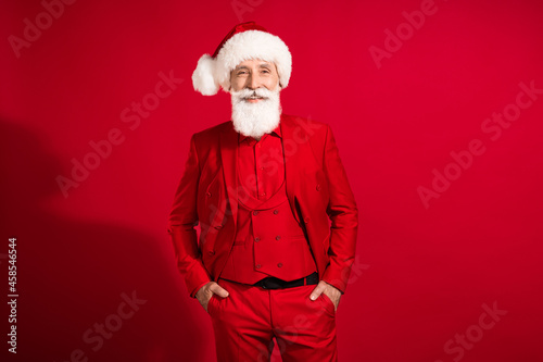 Photo of business man santa claus hands pockets white smile wear x-mas hat tuxedo on red color background