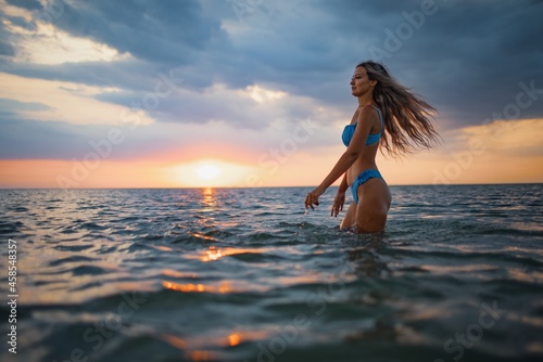 A girl with blond hair in a blue swimsuit splashes to the sides while sitting in an estuary on a sunset background © YouraPechkin