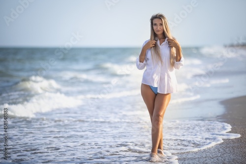 A girl with long hair in a bluish swimsuit and a shirt goes through her hair on the seashore