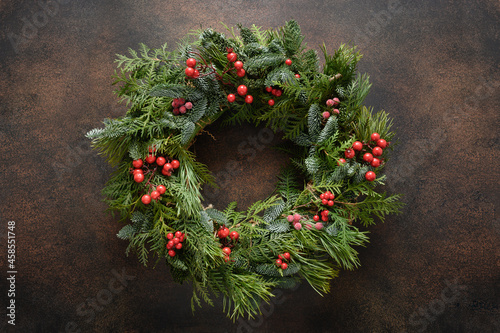 Christmas wreath of natural evergreen branches with red berries on dark brown background. New Year. Traditional decoration for Xmas holiday. View from above. Festive greeting card.
