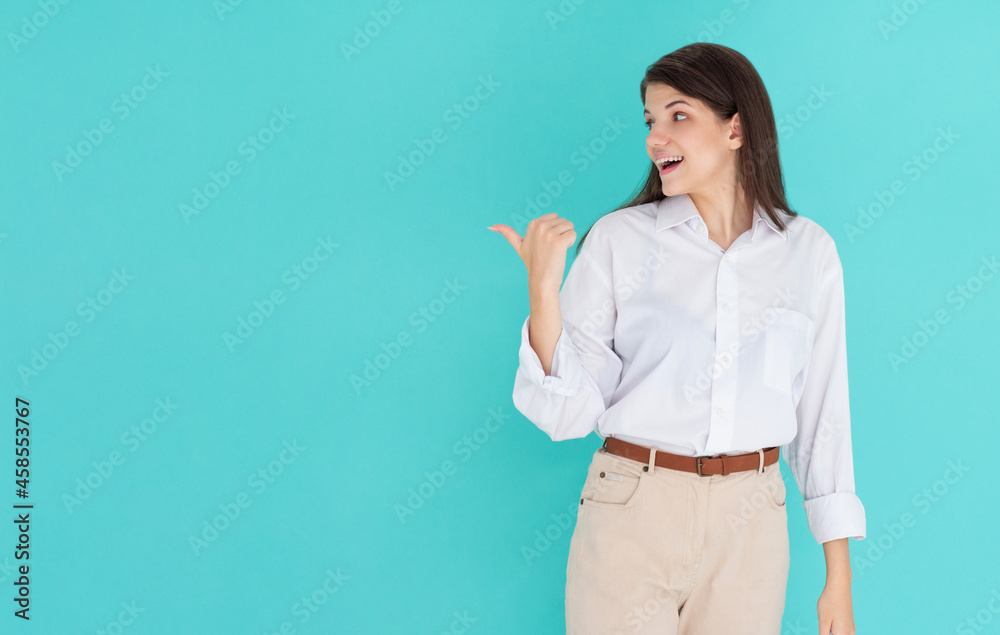 Laughing brunette woman in shirt looking and pointing away with thumb over blue background Laughing brunette woman in shirt looking and pointing away with thumb over blue background