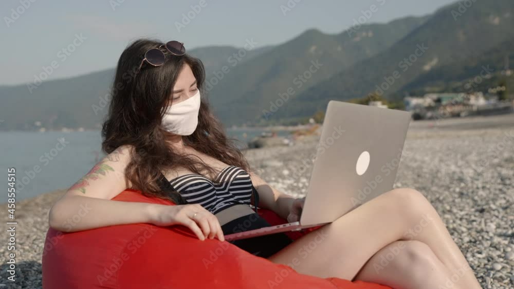 Freelance or distant working during pandemic. Young business woman in bikini in medical mask working on laptop alone by the sea at the beach. Coronavirus protection and social distancing concept. Freelance or distant working during pandemic. Young business woman in bikini in medical mask working on laptop alone by the sea at the beach. Coronavirus protection and social distancing concept.