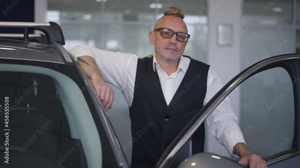 Portrait of successful confident Caucasian businessman in eyeglasses with mohawk haircut posing at open luxurious car in dealership in slow motion. Medium shot of handsome man looking at camera