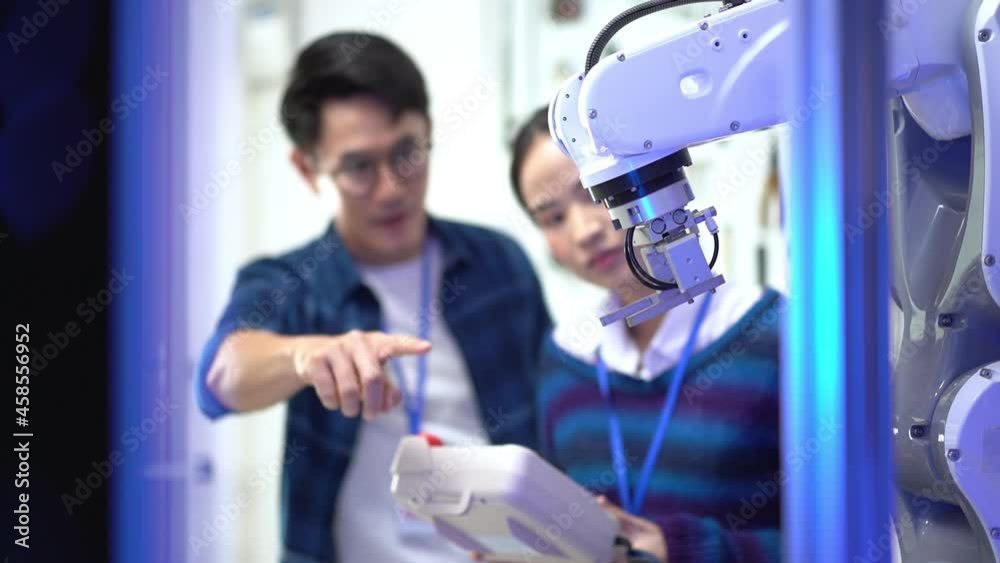 Asian female apprentice engineer in student uniform testing the work of an automation robotic arm machine with a robot controller taught by a young Asian male CEO standing next to her in the factory.