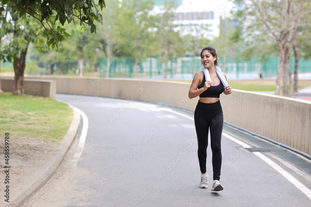 Fototapeta premium Portrait of sporty young asian woman in sportswear jogging outdoor for marathon training. Jogger girl exercising along concrete path outdoor with green tree background. Sport concept