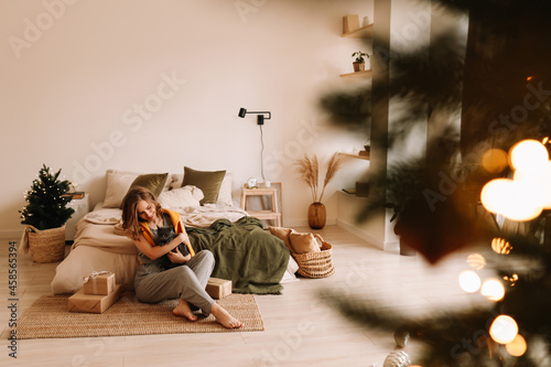 A pretty smiling young woman in pajamas hugs her friend's pet domestic cat sitting in a cozy bedroom decorated with Christmas decor at home, selective focus