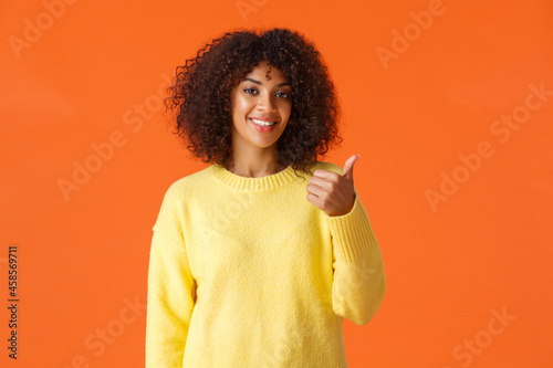 You should come and see it. Carefree good-looking hipster girl yellow sweater, inviting come inside pointing thumb behind herself and smiling, recommend product or company service, orange background