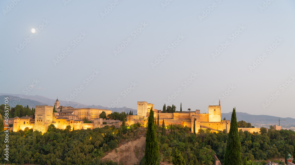 Landscape view of the famous Alhambra illuminated at night, Granada, Andalusia, Spain. White moon on the left. Pale blue sky on the background. With copy-space.