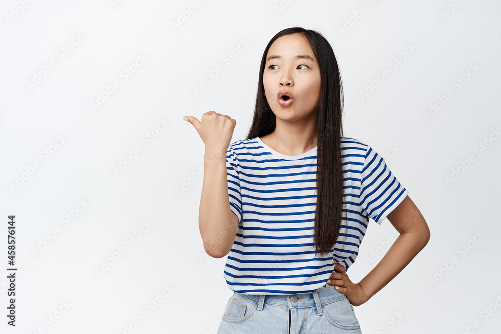 Image of curious asian woman pointing finger left and looking surprised, intrigued by something, showing advertisement, white background Image of curious asian woman pointing finger left and looking surprised, intrigued by something, showing advertisement, white background