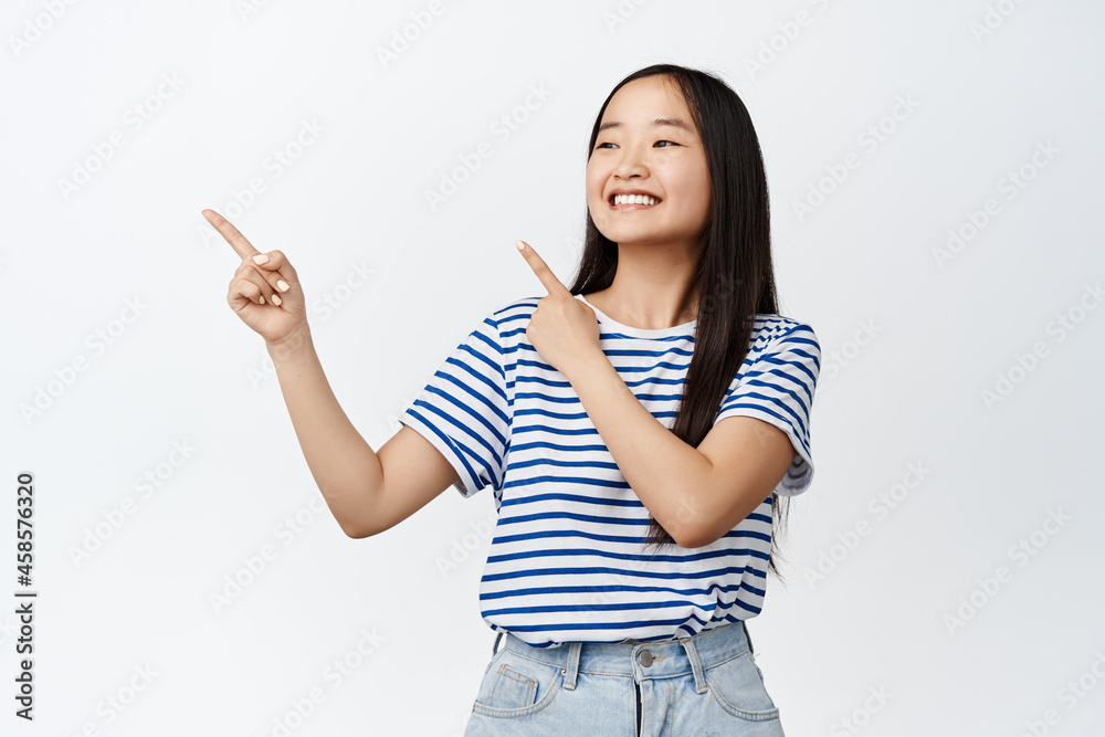 Happy chinese girl pointing fingers and looking left at advertisement, smiling pleased, showing promotion or store logo, standing over white background Happy chinese girl pointing fingers and looking left at advertisement, smiling pleased, showing promotion or store logo, standing over white background