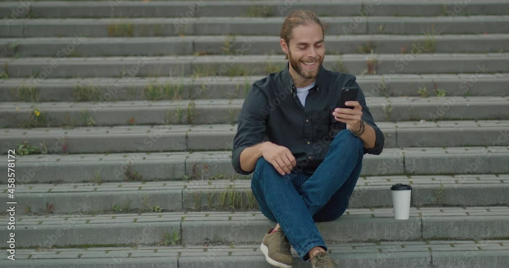 Casual Happy Man Manager, Worker sitting on the stairs using modern smartphone. Handsome Millennial Young Adult Guy spending time surfing on Social Media, Chatting. Messenger. Apps. Casual Happy Man Manager, Worker sitting on the stairs using modern smartphone. Handsome Millennial Young Adult Guy spending time surfing on Social Media, Chatting. Messenger. Apps.