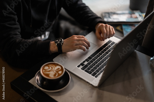 professional man in a black hoodie with a luxury works for hours at a laptop and drinks cappuccino coffee in a cafe. Freelancer guy working indoors, close-up