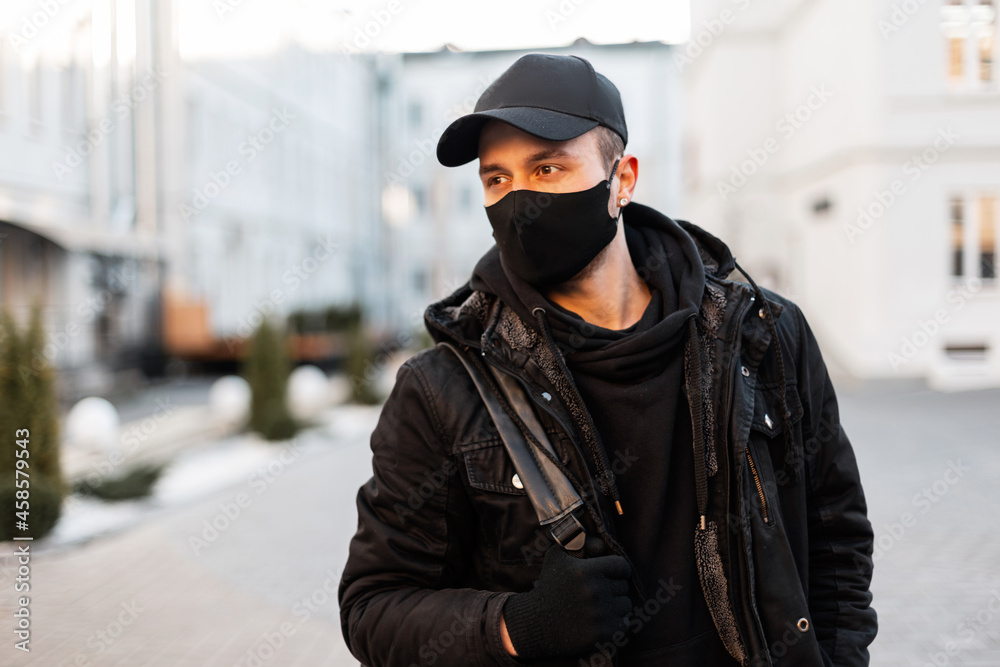 fashionable handsome hipster man with a black cap and an inter-military protective mask in a black jacket, hoodie with backpack walks in the city. Male modern urban style and pandemic concept fashionable handsome hipster man with a black cap and an inter-military protective mask in a black jacket, hoodie with backpack walks in the city. Male modern urban style and pandemic concept