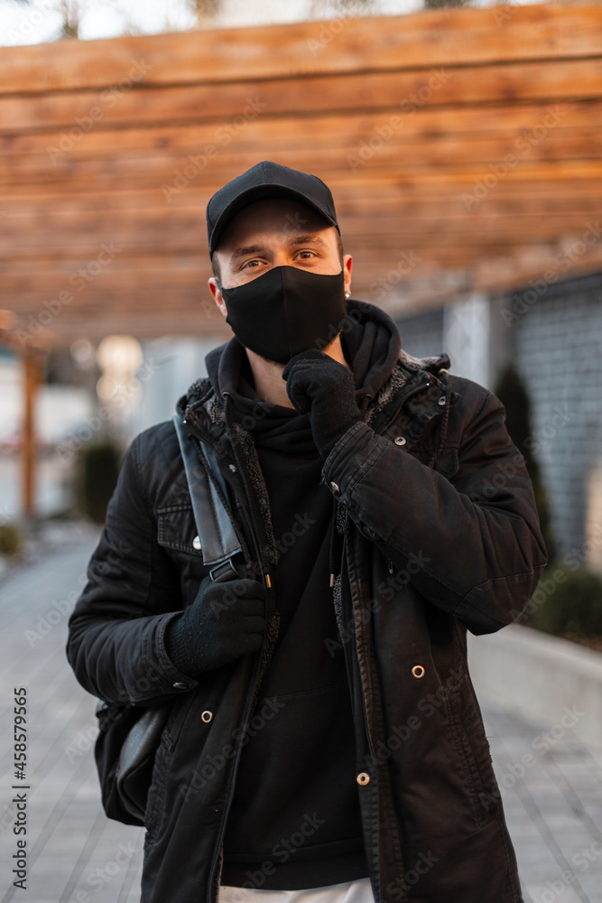 stylish handsome young man with a medical mask and a black cap in a black fashionable jacket, a hoodie with a backpack walks in the city. stylish handsome young man with a medical mask and a black cap in a black fashionable jacket, a hoodie with a backpack walks in the city.