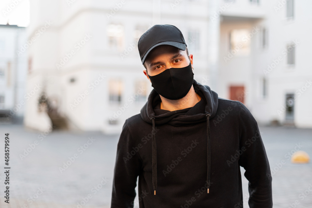Portrait of a handsome young man with a protective black medical mask and stylish cap in a black fashionable hoodie in the city. modern male casual style and coronavirus concept