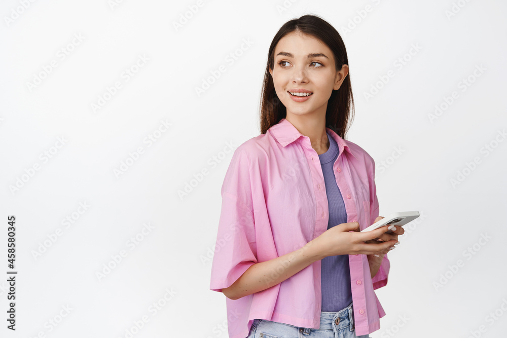 Young modern girl looking aside, holding smartphone, using application, order taxi or chatting on mobile phone app, white background Young modern girl looking aside, holding smartphone, using application, order taxi or chatting on mobile phone app, white background