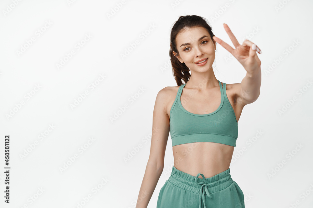 Young fit female athlete showing peace sign. Fitness girl motivating go to gym, workout in activewear, standing over white background Young fit female athlete showing peace sign. Fitness girl motivating go to gym, workout in activewear, standing over white background