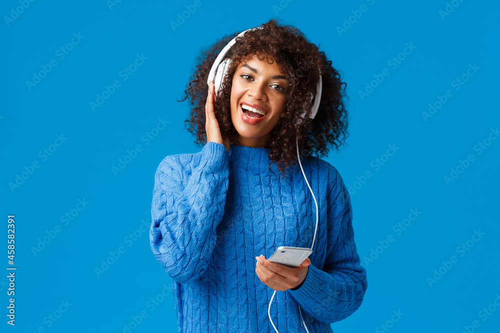 Happy and cheerful african-american woman playing karaoke game app in  smartphone, wearing headphones singing and enjoying awesome music, sound  quality, standing blue background Stock Photo | Adobe Stock