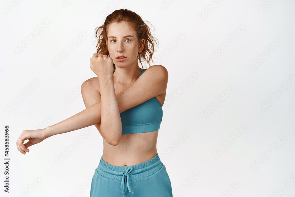 Young female athlete doing sport workout, stretching arm during warm-up, standing in activewear against white background Young female athlete doing sport workout, stretching arm during warm-up, standing in activewear against white background