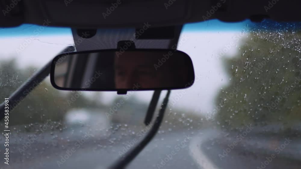 Reflection of middle-aged man in the mirror inside the driving car while heavy raining, rear view of driver holding steering wheel and talking with unrecognisable passenger during road trip, driving