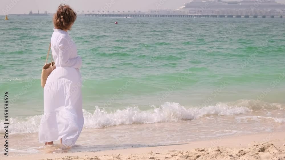 Young white dressed woman is standing on the sandy coast close to sea and looking for horizon. Green water of the ocean with blue waves washes the beach. Nature landscape with lady and copy space. Young white dressed woman is standing on the sandy coast close to sea and looking for horizon. Green water of the ocean with blue waves washes the beach. Nature landscape with lady and copy space.
