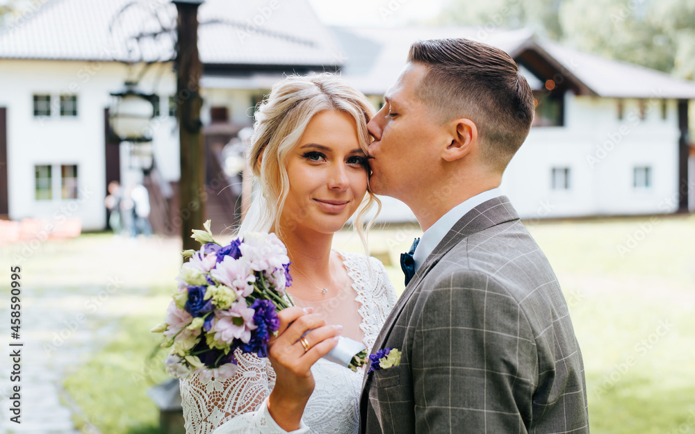 Portrait of young married couple, newlyweds together outdoors, groom kisses forehead of bride in white dress with bouquet flowers. Beautiful happy bride looking at camera. Wedding day, just married