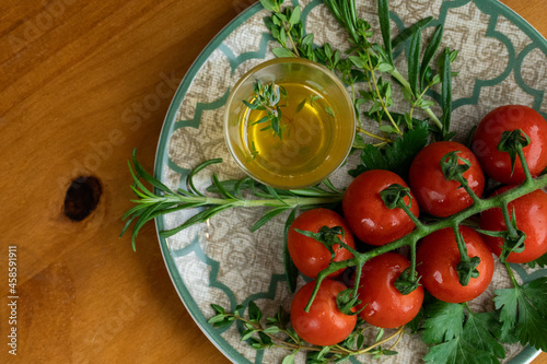 red tomatoes, accompanied by salad, rosemary and thyme, herb oil on the side, served in a plate with green details on a table with wooden background, close-up photo with space for text