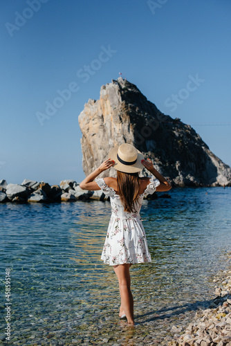 A beautiful young woman in a hat and a light dress with her back is walking along the ocean shore against the background of huge rocks on a sunny day. Tourism and vacation travel. © Andrii