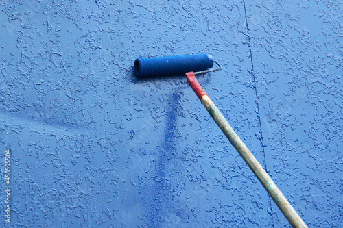 A paint roller on a long handle applies new blue paint to the old one. Construction and renovation concept.