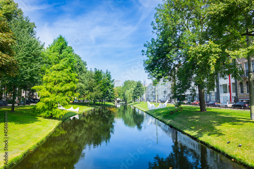 Urban park leafy trees idyllically reflected in calm water of canal running through city under large leafy tree