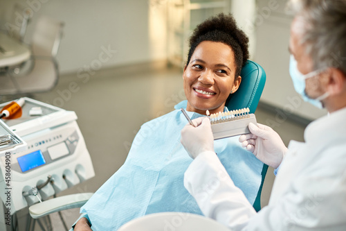Happy black woman and her dentist choosing the shade of dental veneers during appointment at dental clinic.