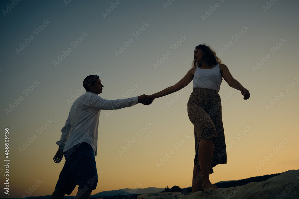 Young adult man and woman holding hands looking to each other against seaside sunset. Woman leads man after her holding his hand