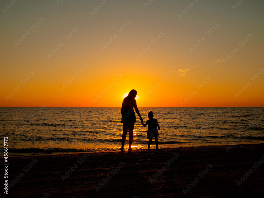 A little girl holds her mother's hand, standing on the sea beach on a summer evening. In the background, the evening sun setting over the horizon.