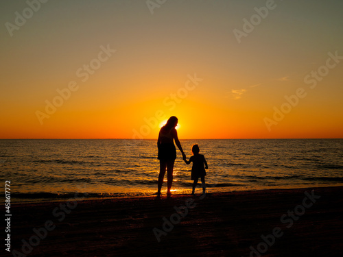 A little girl holds her mother's hand, standing on the sea beach on a summer evening. In the background, the evening sun setting over the horizon.