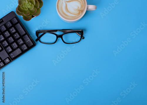 cup of coffee, glasses,  plant, black keyboard on blue background top view Space for text