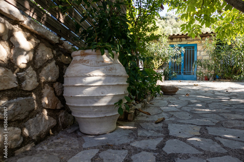 Big flower vase in green sunny summer in Greek traditional yard with stone walls and blue doors shutters. Summer travel locations architecture details © Kathrine Andi