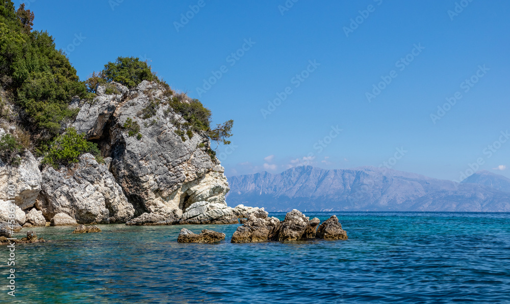 Scenic rocks in blue water of Ionian Sea with green cliffs, distant mountains and bright sky. Nature of Ormos Desimi, Lefkada island in Greece. Summer vacation travel destination