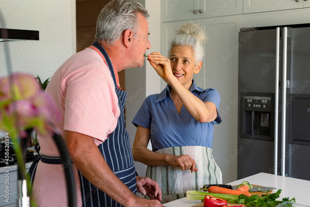 Fototapeta premium Happy caucasian senior couple standing in kitchen, preparing meal together and eating vegetables