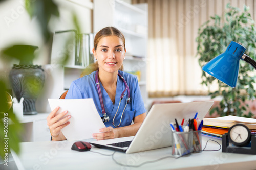 Female doctor in surgical scrubs sitting at desk in office and looking in documents in her hands.