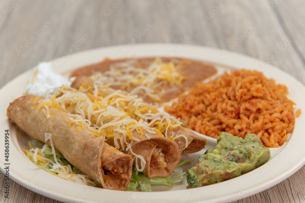 Taquitos rancheros, covered in shredded cheese and served on a plate with guacamole, rice, and beans for a very hearty Mexican meal Taquitos rancheros, covered in shredded cheese and served on a plate with guacamole, rice, and beans for a very hearty Mexican meal