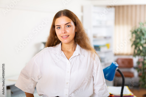 Free Download Closeup portrait of positive brown-haired young woman wearing white blouse posing on blurred background of modern office interior - GFXLoad
