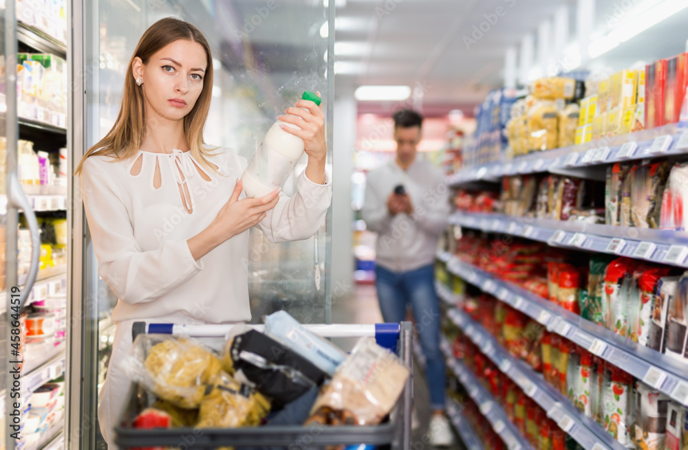 Portrait of young happy positive smiling woman with shopping cart choosing dairy products in supermarket
