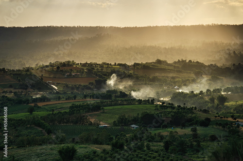 Sunset over the mountains in cloudy sky with sun rays down to the hills with green forest and agricultural field