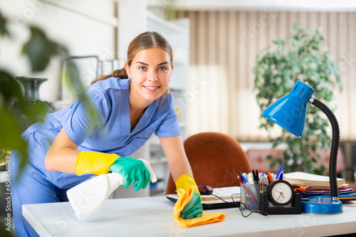 Obraz na plátně Portrait of a positive young female worker cleaning an office desk with a rag an
