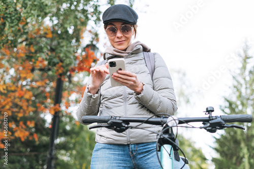 Fototapeta Naklejka Na Ścianę i Meble -  Portrait of fashionable young pretty woman in cap and sunglasses using mobile on bicycle on autumn day in city park